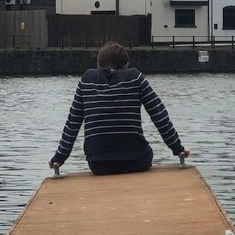 A photograph of a man sat at the end of a small jetty, facing away from the camera