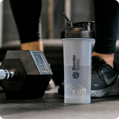 A bottle from BlenderBottle on a gym floor, next to a set of dumbbells, in front of an athlete.