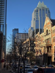 A sunny city street featuring a mix of historic architecture and modern glass skyscrapers under a clear blue sky, with parked cars and leafless trees lining the sidewalk.