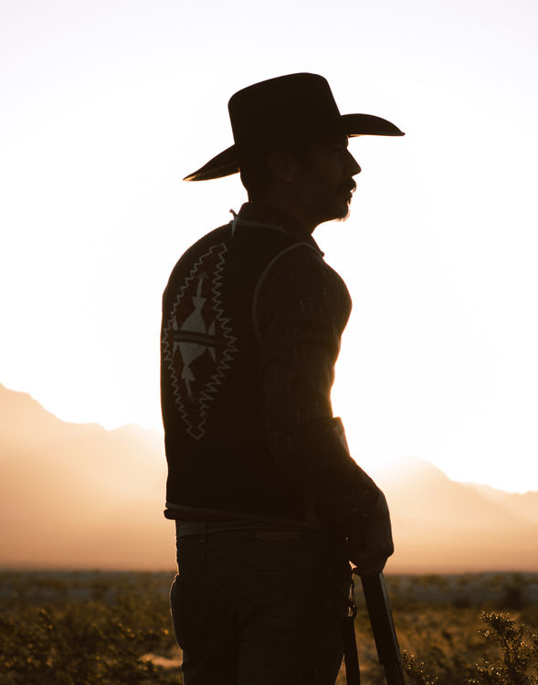 Desert cowboy portrait session at golden hour by Daniela Blagoeva, specializing in Western lifestyle, portrait, and boudoir photography. Air Force veteran in a black hat and textured leather vest, standing under warm sunset light with a soft desert backdrop.