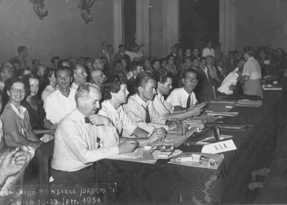 A group of men and women sit at tables in a conference room in 1954. A sign labeled "USA" is visible on the table.