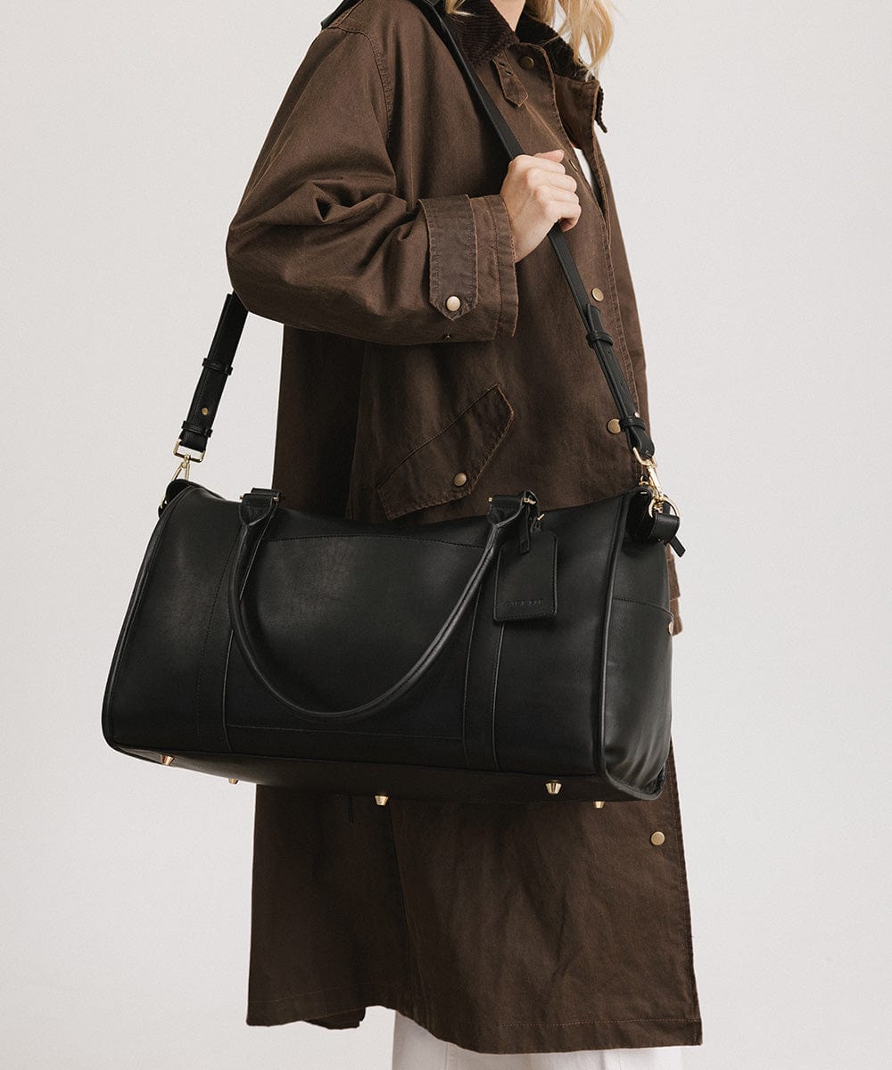 Person wearing a brown coat carrying a black vegan leather weekender bag against a plain background #color_black